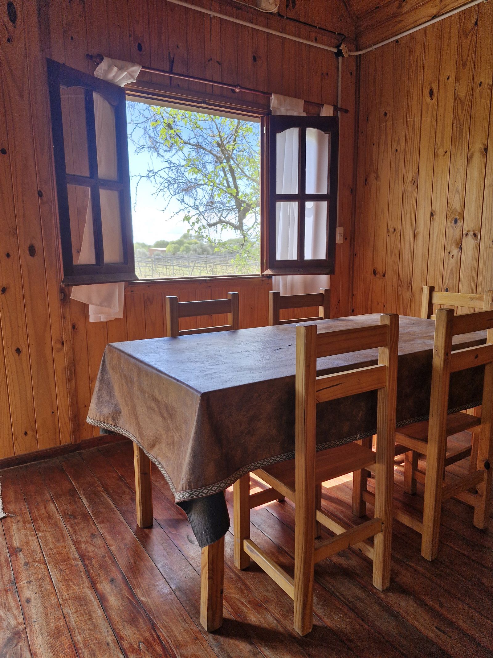 Mesa de comedor con sillas de madera junto a una ventana abierta que da al viñedo