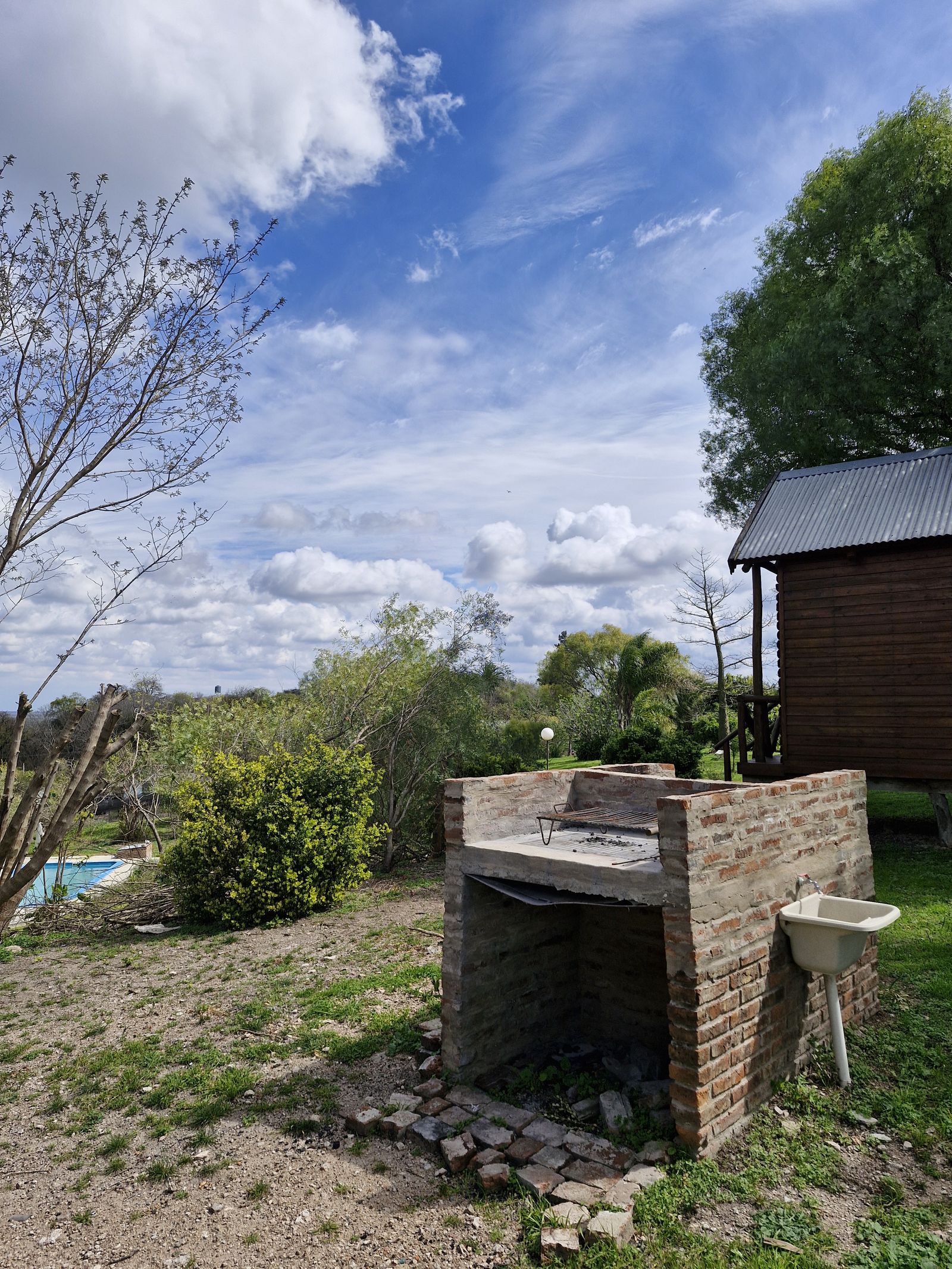 Parrillero de ladrillo con pileta de lavado; al fondo, la piscina y una cabaña